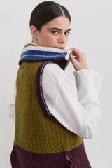 Chaleco de lana con cierre frontal completo y cuello alto. Presenta un diseño de bloques de color en tonos marrón chocolate y celeste, combinando secciones de tejido trenzado con secciones de tejido acanalado. Posee un corte asimétrico, siendo más largo en la parte posterior, y aberturas laterales en el ruedo.