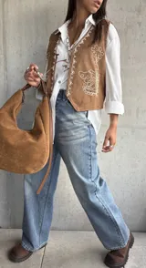 Chaleco de gamuza color camel con bordados en hilo blanco y detalles de tachas metálicas. Presenta un diseño abierto con terminaciones redondeadas en el frente.