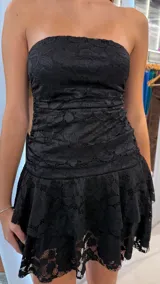 Vestido corto de encaje negro con diseño strapless, cuerpo drapeado y falda con volados en capas.