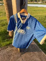 Camiseta tipo polo de fútbol Scuffers color azul, de corte boxy fit, confeccionada en tejido microperforado. Presenta cuello y puños en color crudo, franjas verticales blancas en el lado derecho y estampados tipográficos en el pecho.