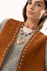 Chaleco oversize confeccionado en cuero descarne, con diseño de terminación en picos, detalle de tachas metálicas en los bordes frontales y flecos en la espalda.