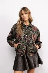 Campera tipo bomber con diseño de estampado floral estilo tapicería sobre fondo negro. Presenta cuello clásico, cierre frontal con botones metálicos, bolsillos laterales y puños elásticos.