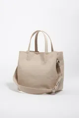Bolso de mano grande tipo tote de Bimba y Lola, color beige, con doble asa superior y correa ajustable de tejido. Presenta logo metálico en la parte inferior y cierre superior.