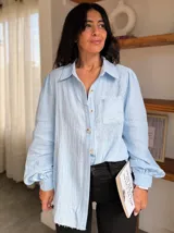 Camisa de tela texturizada color celeste, con mangas largas abullonadas, cuello clásico y cierre frontal con botones. Presenta un delicado bordado floral en la parte superior delantera y un bolsillo lateral.