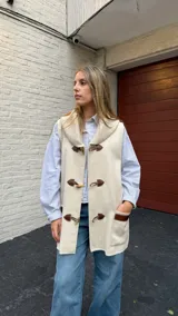 Chaleco de lana merino color beige, con cuello tipo chal acanalado y cierre frontal mediante tres trabillas de cuero con botones tipo duffle. Presenta dos bolsillos delanteros con detalles en cuero y un diseño de corte recto y largo.