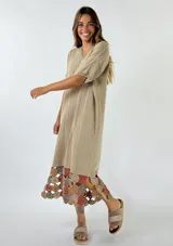 Vestido midi de tejido de punto color beige, con corte holgado y mangas cortas. Presenta un diseño con terminación en el ruedo de aplicaciones circulares tejidas a crochet en tonos tierra y rosado.