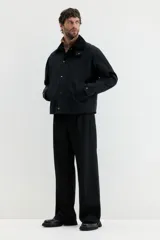 Campera negra de sarga encerada de algodón con cuello de pana. Presenta cierre frontal cubierto por una solapa con botones a presión, mangas raglán con puños ajustables mediante botones a presión, bolsillos insertados al bies y un bolsillo interior con botón a presión. Tiene un corte holgado y forro interior.