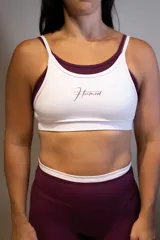 Top deportivo de entrenamiento con doble capa, color blanco exterior y borgoña interior. Presenta un corte de tirantes anchos y el logo "Hermod" en cursiva color borgoña en el pecho.