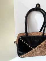 Cartera rígida negra con detalles en rojo y tachas plateadas. Tiene herrajes y cierre metálico plateado marca YKK, asa fina para llevar al hombro o en la mano y correa larga extra regulable. El interior es forrado, tiene un bolsillo con cierre enmarcado en cuero, y un segundo bolsillo plaqué.