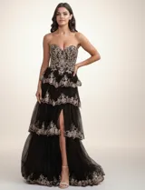 Vestido largo de fiesta con escote corazón y corset estructurado. Presenta un diseño de capas de tul con volados y bordados brillantes en tonos dorados sobre base negra. Incluye un tajo lateral profundo en la falda.