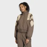 Campera deportiva Adidas de corte holgado, con cierre frontal y cuello alto. Presenta un diseño en bloques de color marrón y beige, con detalles inspirados en la estética futbolística de los años 90.