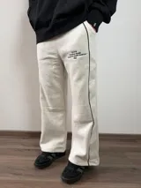 Pantalón tipo jogger de corte baggy, confeccionado en algodón color gris melange. Presenta una línea vertical negra a lo largo de la pierna y un estampado tipográfico pequeño en la parte superior frontal.