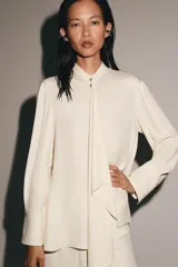 Blusa de viscosa color marfil, con cuello mao, manga larga con puños anchos, cierre frontal con botones y detalle de lazada colgante en el frente.