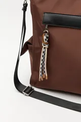 Bolso tipo tote color negro, con doble asa superior y correa desmontable. Presenta un bolsillo frontal con solapa, bolsillos laterales y un detalle colgante decorativo con tiras de colores.