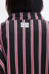 Campera de denim oversize con cierre frontal, cuello alto y bolsillos delanteros. Presenta un diseño a rayas verticales en tonos negros y rosados, cruzado por una franja ancha horizontal de color blanco con forma de zigzag.