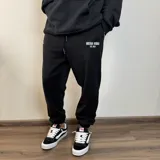 Pantalón tipo jogger de algodón color negro, con cintura elástica ajustable mediante cordón y puños elásticos en los tobillos. Presenta un estampado pequeño en la parte frontal superior con la frase 'BUENA VIBRA EST. 2018'.