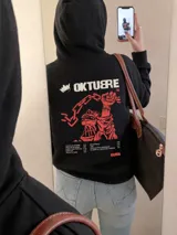 Canguro negro de corte regular con capucha y bolsillo frontal. Presenta un estampado gráfico en la espalda en color rojo y crema que incluye una mano sosteniendo una cadena, el título 'Oktubre' y un listado de canciones.
