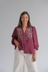 Blusa color bordó con bordados florales en tono crema en la parte superior frontal. Presenta cuello redondo con pequeña abertura, cierre frontal con botones y mangas largas acampanadas con puños ajustados.