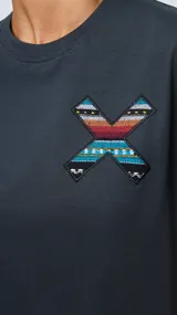 Remera blanca de algodón orgánico con cuello redondo y manga corta. Presenta un estampado en el pecho con una cruz formada por dos tablas de surf en tonos rojo, azul y negro.