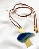 Collar con dije de ágata azul irregular sobre una placa rectangular de latón. El collar tiene un cordón de cuero marrón con cuentas doradas en los extremos. El dije mide 6,5 x 3 cm y el engarce mide 2 x 6,5 cm. El cordón mide 140 cm de largo.