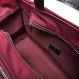 Bandolera de tela color bordó con detalles en negro, cuenta con doble asa de mano, correa ajustable extraíble, cierre principal y bolsillo frontal con cierre, además de un detalle de cordón decorativo y logo circular metálico en el frente.
