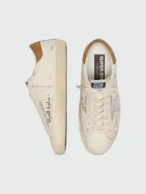 Zapatillas Golden Goose modelo Super-Star de cuero napa color blanco, con la icónica estrella lateral laminada en plateado, talón de gamuza marrón y efecto desgastado artesanal. Presenta inscripciones y detalles escritos a mano en los laterales y la lengüeta.