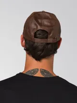 Gorro tipo gorra de béisbol con visera curva, confeccionado en cuero sintético con acabado texturizado. Presenta un pequeño aplique metálico con el logo de la marca en el lateral frontal y cierre metálico ajustable en la parte posterior.