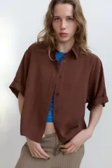 Camisa de cuello solapa y manga corta con dobladillo. Presenta un diseño de corte holgado con bajo asimétrico y aberturas laterales. Cierre frontal mediante botones ocultos bajo una tapeta.