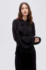 Camisa negra de poliéster, de corte holgado, con cuello redondo y lazo para anudar al frente, y mangas largas abullonadas con puños ajustados.