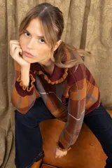 Campera de punto con diseño a cuadros tartán en tonos marrones y beige. Presenta un corte oversize, cierre frontal con cremallera, cuello tipo babero con volados y mangas largas con puños ajustados.