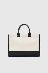 Tote bag de diseño estructurado, confeccionado en lona color crema con base, asas y detalles en cuero negro. Presenta un logo circular en relieve en el frente y correa larga ajustable para llevar al hombro.