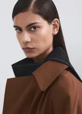 Chaqueta larga tipo trench color marrón, con diseño recto, solapas anchas, doble botonadura, bolsillos laterales y cinturón desmontable de la misma tela.