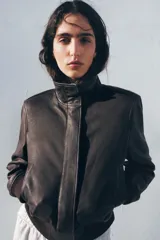 Chaqueta tipo bomber de piel con cuello alto, manga larga con hombreras y puños elásticos. Presenta bolsillos delanteros, bajo elástico y cierre frontal con cremallera y botones ocultos por solapa.