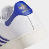 Championes Adidas Jabbar Lo de diseño clásico inspirado en los años 70. Presentan una parte superior de cuero blanco con las icónicas tres tiras laterales en azul, detalle en el talón a juego y puntera de gamuza gris claro. Cuentan con suela de goma y mediasuela de EVA para mayor comodidad.