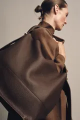 Bolso tipo saco de gran tamaño en cuero marrón oscuro, con textura granulada. Presenta un asa de hombro ancha y detalles metálicos dorados en la unión del asa. Incluye un bolsillo interior extraíble y cierre mediante botón imantado.