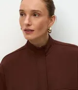 Campera de corte recto y largo, color marrón chocolate, con cuello alto, cierre frontal completo y bolsillos frontales tipo cargo con solapa. Presenta puños y ruedo acanalados.