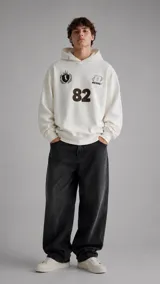 Hoodie de algodón color crema con capucha, corte holgado y diseño estampado en el frente que incluye el número 82, un escudo heráldico y el logo de la marca Duxon.