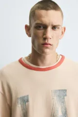 Camiseta de punto tejida color beige claro, corte boxy fit, con cuello redondo y mangas cortas. Presenta estampado desgastado con las letras "LA" en el frente y ribetes en color terracota en cuello y mangas. El bajo es irregular.