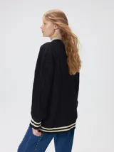 Cardigan de tejido de punto con diseño de ochos, corte oversize y mangas largas con puños acanalados. Presenta un escote en V con solapas en contraste, cierre frontal con botones y dos bolsillos delanteros con bordes en contraste. El diseño incluye detalles de rayas en los puños y en el ruedo.