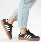 Championes Adidas modelo Sambae, color negro con las icónicas tres tiras blancas laterales y suela de goma color caramelo con plataforma. Presenta detalles en cuero y refuerzo de gamuza en la puntera.