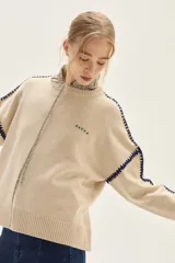 Sweater de tejido de punto color beige, con cuello redondo y detalles de bordado en zigzag en color azul oscuro a lo largo de las mangas y hombros. Presenta un pequeño bordado con texto en el pecho.