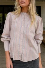 Camisa de algodón a rayas en tonos rosado, celeste y gris, con cuello mao, cierre frontal con botones, pliegues decorativos en el pecho y mangas abullonadas con puños largos.