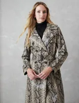 Trench de ecocuero con estampado animal print de serpiente en tonos beige, marrón y negro. Presenta cuello con solapa ancha, doble abotonadura frontal con botones negros, cinturón ajustable con hebilla metálica, mangas largas con presillas y botones en los puños, canesú suelto en la espalda, bolsillos laterales y forro interno.