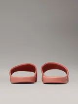 Ojotas tipo slide de color rosado, con base de EVA y correa ancha de lona de algodón reciclado. La correa presenta el logo de Calvin Klein Jeans bordado en blanco.