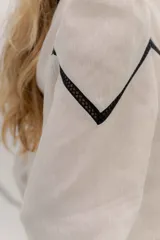 Camisa de lino color beige con mangas abullonadas y detalles de puntilla negra en forma de V en el frente. Presenta cuello a la base con borde desflecado, cierre frontal con botones forrados y puños con alforzas.