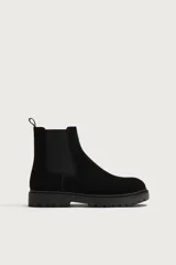 Bota tipo Chelsea de hombre en color negro, confeccionada en cuero con acabado de serraje. Cuenta con elásticos laterales, tirador trasero y suela dentada con volumen de estilo track.