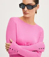 Suéter de punto color rosado, con cuello barco, mangas largas y puños con botones decorativos. El dobladillo también es acanalado.