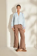 Blusa de lino color marrón con escote en V, cuello con volados y mangas largas con puños fruncidos que presentan detalles en color crudo, al igual que en los hombros. Cuenta con cierre frontal de botones.