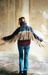 Campera de lana italiana con diseño de motivos tribales en tonos oscuros, rojos y beige. Presenta cierre frontal, bolsillos laterales y detalle de flecos largos de gamuza en las mangas.