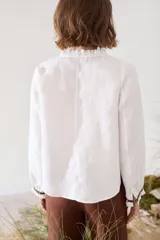 Blusa de lino color marrón con escote en V, cuello con volados y mangas largas con puños fruncidos que presentan detalles en color crudo, al igual que en los hombros. Cuenta con cierre frontal de botones.